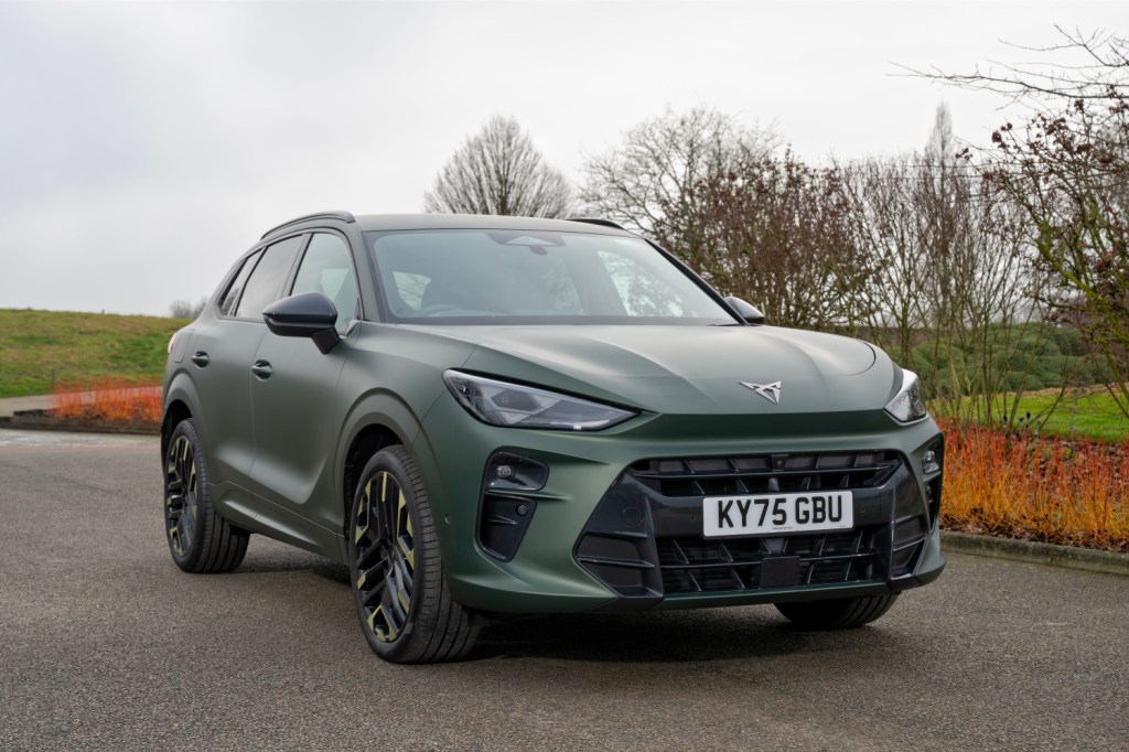 A sleek, green SUV parked on a road with a grassy background and bare trees.