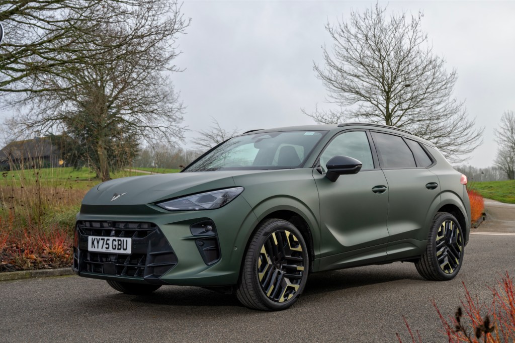 A sleek, green SUV parked on a road, showcasing modern design and stylish black and gold wheels, with trees and grass in the background.