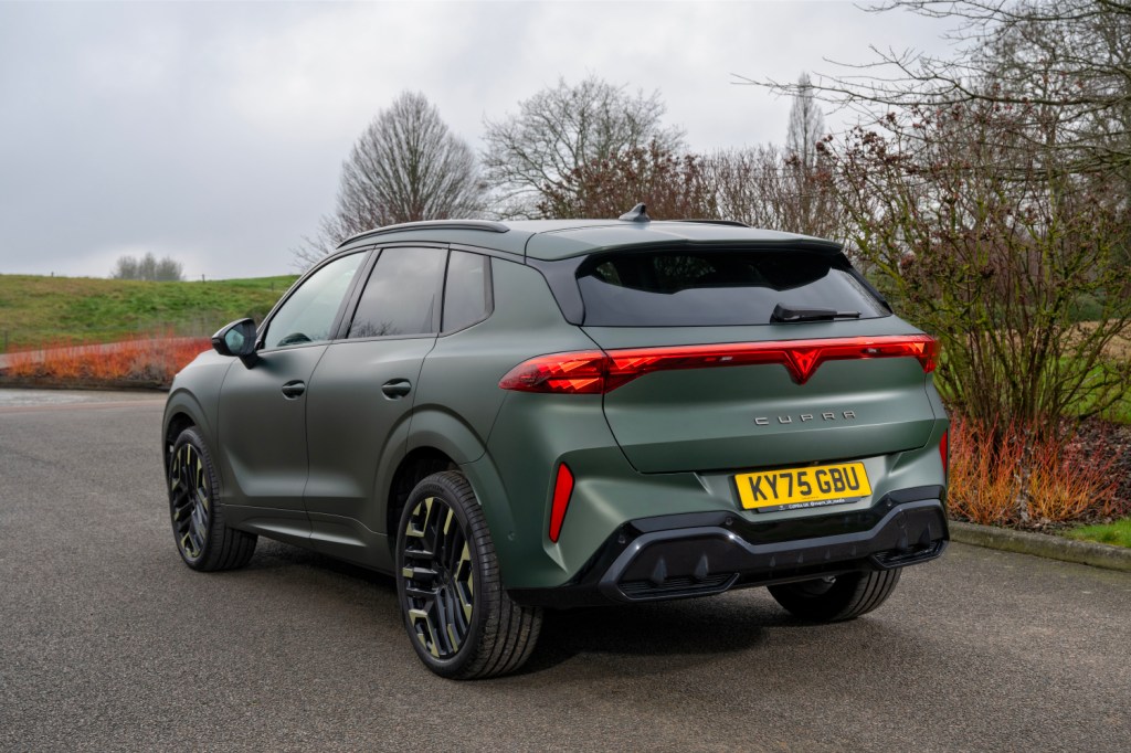 A rear view of a green Cupra SUV parked on a road, showcasing its sleek design, distinctive taillights, and stylish alloy wheels, with a backdrop of trees and cloudy sky.